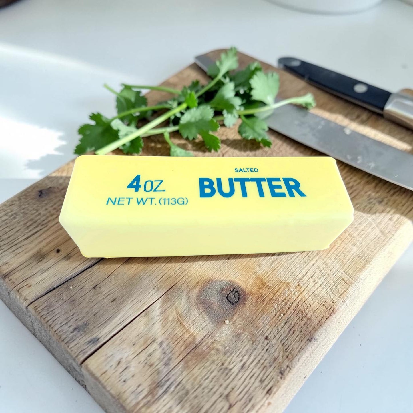 Block of butter squishy on a wooden cutting board with herbs and a knife in the background.