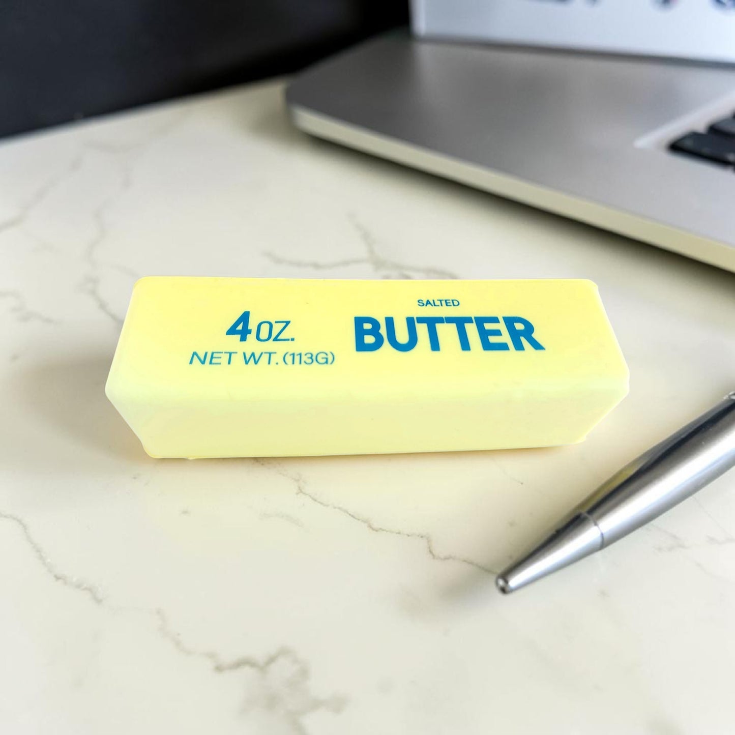 butter squishy, Bar of butter labeled '4 oz Salted Butter' on a marble surface with a laptop in the background.
