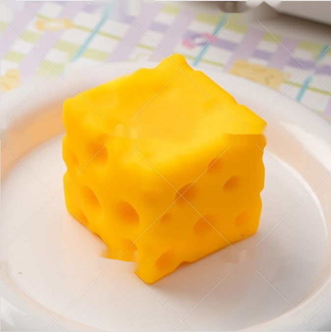 cheese squishy, Yellow cheese cube-shaped object on a white plate with a checkered tablecloth in the background