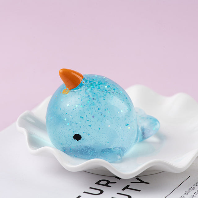Blue color Mochi Squishy, Blue glittery whale figurine with an orange horn on a white shell against a light purple background.